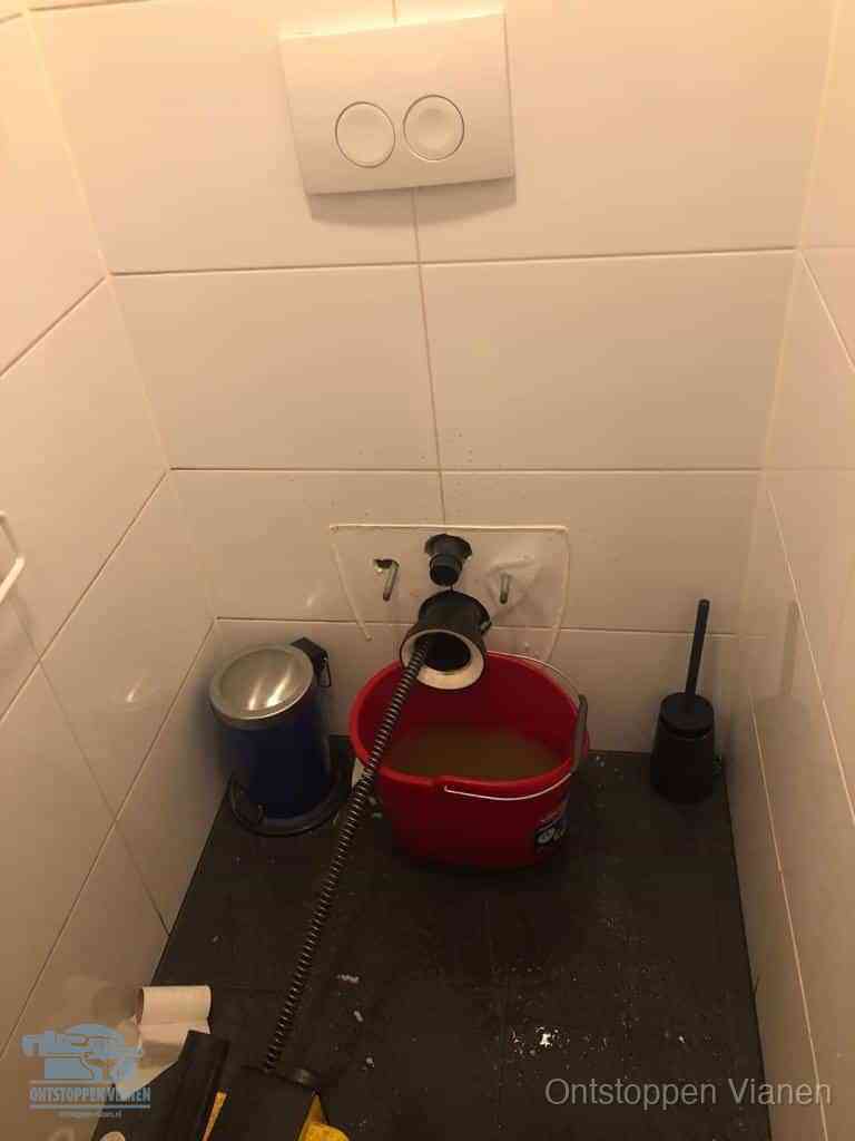 Een ontstopte wand-wc met verwijderde toiletpot waar een ontstoppingsveer in de wanduitgang verdwijnt, het achterhalen waar de verstopping zit in Vianen