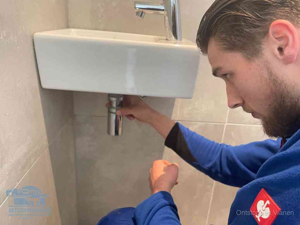 Een monteur draait met de hand de zwanenhalssifon onder een fonteinwasbak in de toiletruimte los om de bron van rioollucht in Vianen te vinden