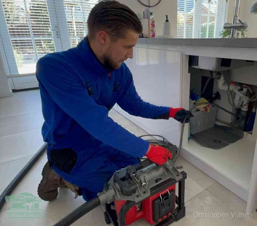 Een monteur met rode handschoenen knielt in een keuken en voert een elektrische ontstoppingsveer in tijdens het professioneel afvoer ontstoppen in Vianen